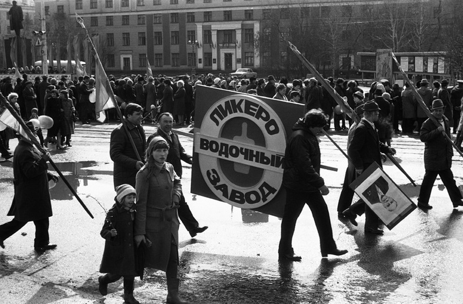 Возвращение с первомайской демонстрации - торжественного прохождения колонн трудящихся перед руководителями района, стоящими на трибуне. Праздник Международной солидарности трудящихся всех стран. В руках бывших демонстрантов свернутые флаги, портреты руководителей государства и тяжелый стенд с названием завода. Площадь Ленина. Кузнецкий район. Новокузнецк. Кемеровская область. Кузбасс. Сибирь. 1.05.1983.