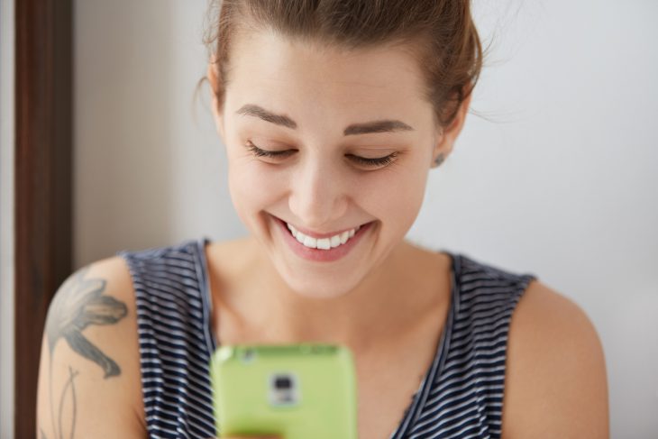 Happy European female chatting in social networks with her friends. Young attractive girl with shiny smile laughing at jokes from messenger, texting back sms to friends with her green digital device.